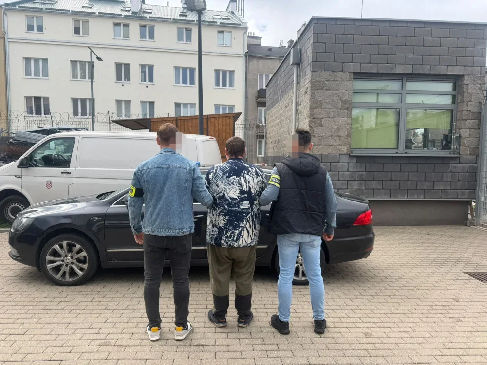 Trzech mężczyzn, z których dwóch nosi opaski identyfikacyjne na ramionach, stoi przy czarnym samochodzie na brukowanym podwórzu przed budynkami.