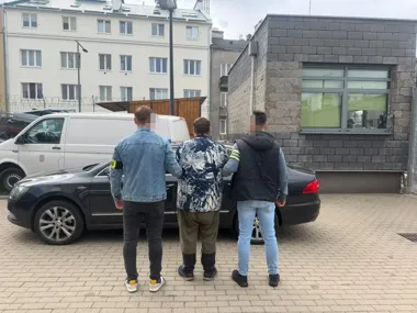 Trzech mężczyzn, z których dwóch nosi opaski identyfikacyjne na ramionach, stoi przy czarnym samochodzie na brukowanym podwórzu przed budynkami.